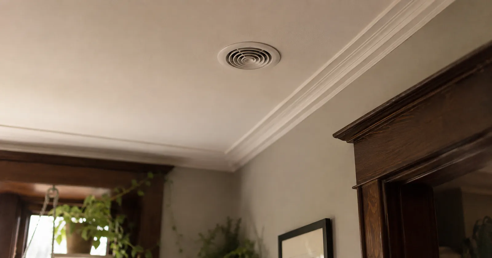Small round white high-velocity air outlet set flush into the ceiling of a heritage room beside crown moulding and dark wood door trim