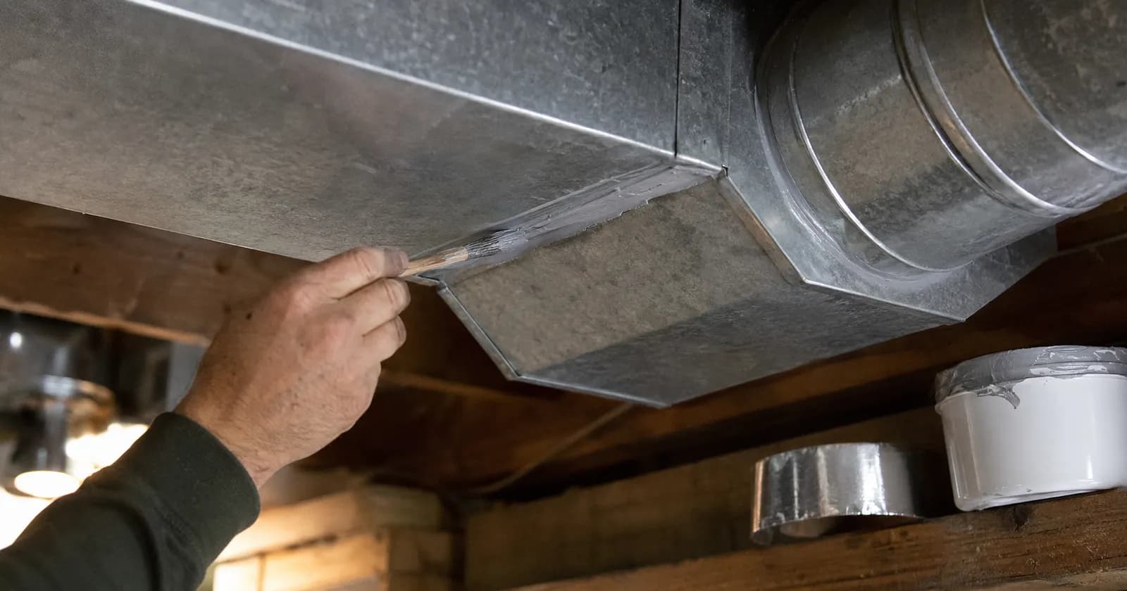 Hand brushing grey mastic sealant onto the seam of a galvanized sheet-metal duct joint in a basement