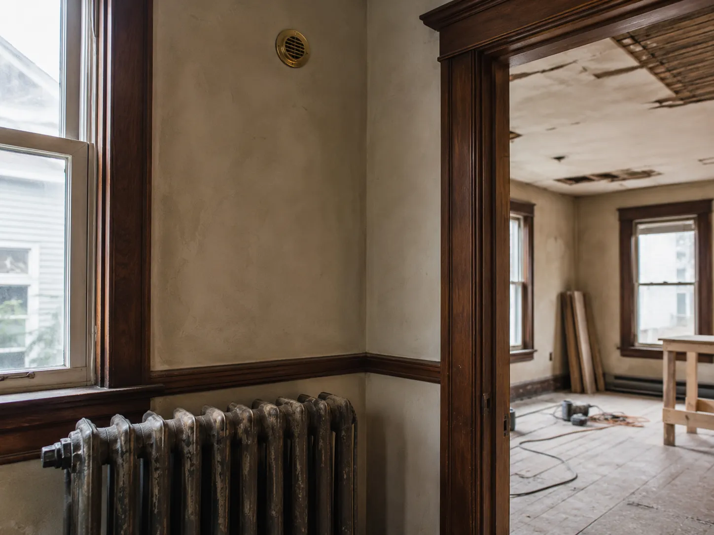 Cast-iron radiator and a small round brass air outlet on the plaster wall of a century home mid-renovation, original dark wood trim throughout